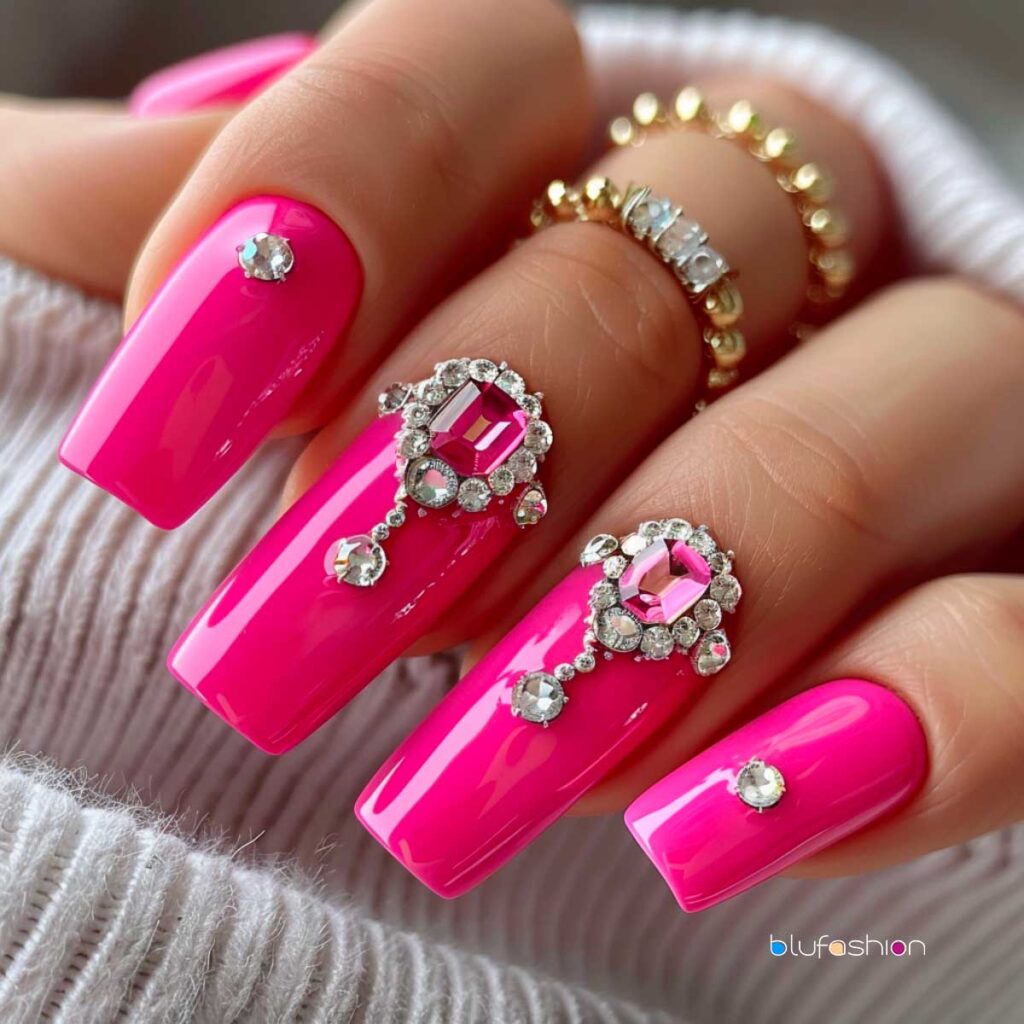 Ongles de cercueil luxueux rose vif ornés de gros strass en diamant rose et clair, créant une manucure élégante et opulente, complétée par des bagues dorées empilées sur les doigts.