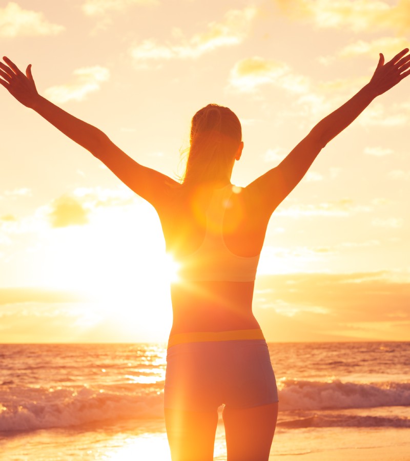 A happy woman rejoices with her arms waving in the air while the sun sets. She